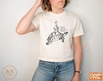 May include: Cream-colored t-shirt featuring a black illustration of a turtle carrying a house on its back. The text "everyone deserves a safe space to call home" is printed above the turtle. The shirt is a casual wear item.