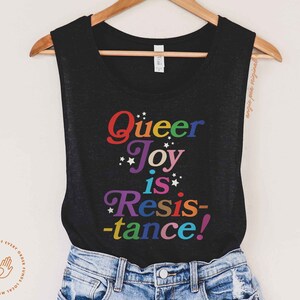 Op de afbeelding: Een zwart tanktop met de tekst "Queer Joy is Resistance!" bedrukt in regenboogkleuren. De tekst is in een speels, retro lettertype.
