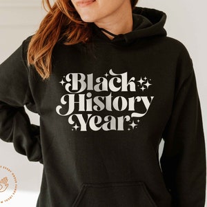 K&ouml;nnte beinhalten: Schwarzer Kapuzenpullover mit dem Text "Black History Year" in wei&szlig;er Retro-Schriftart mit Sternen. Der Hoodie hat eine Vordertasche und eine Kapuze mit Kordelzug.