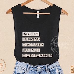 May include: Black sleeveless shirt with the text "IMAGINE FEARING DIVERSITY BUT NOT DICTATORSHIP" in white block letters. The shirt is paired with blue denim shorts, hanging on a wooden hanger.