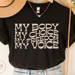 May include: Black t-shirt with the text "MY BODY MY CHOICE MY FREEDOM MY VOICE" in a beige color. The text is in a retro, bold font.