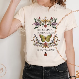 May include: A white t-shirt with a floral design and the text "Decolonize Your Garden" and "Plant Native". The design features a butterfly, a bee, a ladybug, and a crescent moon.