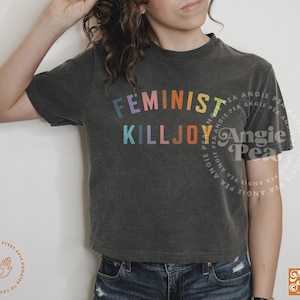 May include: Dark gray t-shirt with the text "FEMINIST KILLJOY" in rainbow colors. The shirt is a cropped style and is paired with blue jeans. The design includes the Angie Pea logo.