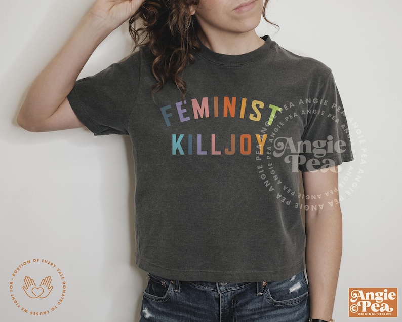 Może przedstawiać: Ciemnoszara koszulka z napisem "FEMINIST KILLJOY" w tęczowych kolorach. Koszulka ma kr&oacute;tki kr&oacute;j i jest połączona z niebieskimi dżinsami. Projekt zawiera logo Angie Pea.