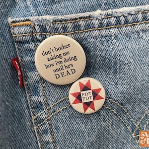 May include: Two round pinback buttons on a denim background. One button has the text "don't bother asking me how I'm doing until he's DEAD". The other button has a starburst design with the letters "FDT".