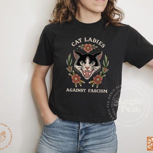 Puede incluir: Ilustraci&oacute;n de un gato blanco y negro con flores y el texto "Cat Ladies Against Fascism" en una camiseta negra.