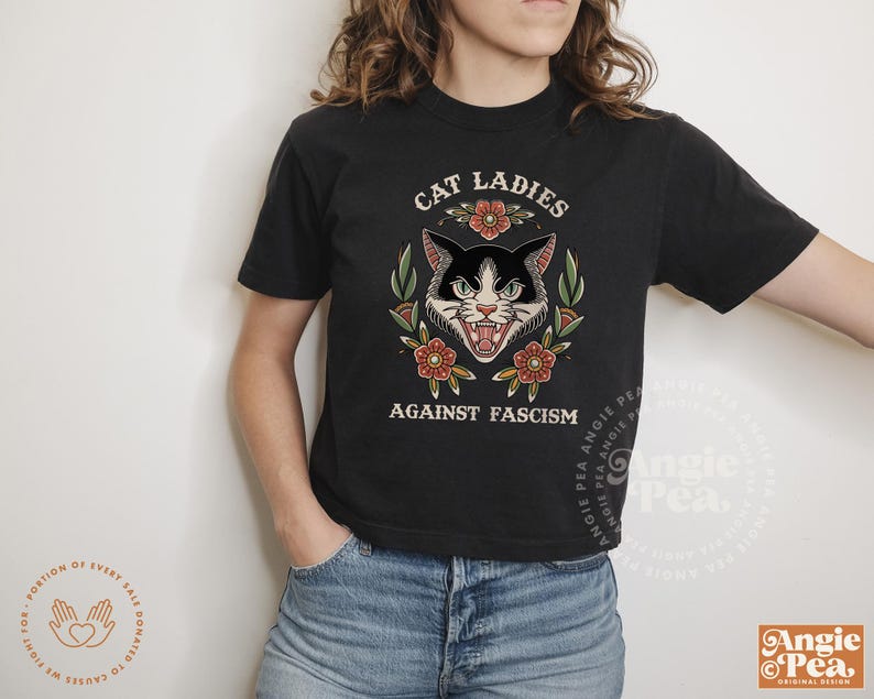 Puede incluir: Ilustraci&oacute;n de un gato blanco y negro con flores y el texto "Cat Ladies Against Fascism" en una camiseta negra.