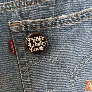 May include: A round, black button with the text "Public Library Lover" in white script is pinned to the back pocket of a pair of blue denim jeans. The jeans have a distressed look with a red Levi's tag visible.