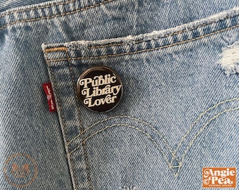 May include: A round, black button with the text "Public Library Lover" in white script is pinned to the back pocket of a pair of blue denim jeans. The jeans have a distressed look with a red Levi's tag visible.