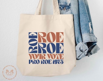 May include: A cream-colored canvas tote bag featuring a graphic print with the text "ROE ROE YOUR VOTE PRO ROE 1973" in blue and terracotta colors. The design uses a bold, stylized font, and the bag has two fabric handles for carrying.