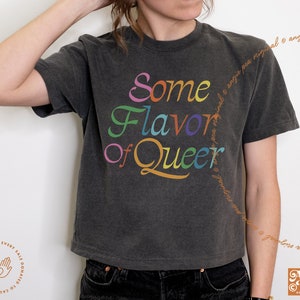 May include: Dark gray t-shirt with the text "Some Flavor of Queer" in rainbow colors. The shirt has a vintage, washed look. The design includes the Angie Pea Original Design logo.
