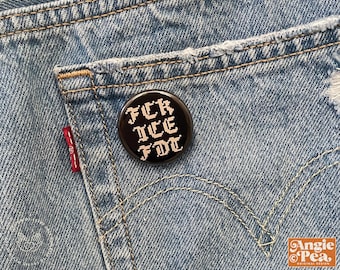 May include: A black button pin with white pixelated text that reads "FCR ICE FDT" is pinned to the back pocket of a pair of blue denim jeans. The jeans have a red tag and distressed details.