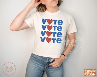 May include: Cream-colored t-shirt with the word "VOTE" in blue letters and red heart symbols. The design is repeated four times down the front of the shirt. The shirt is paired with blue jeans.