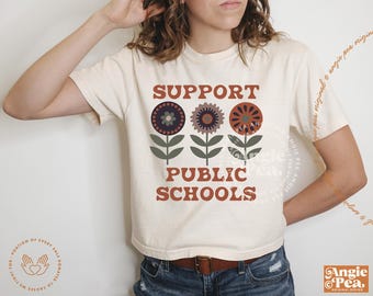 Puede incluir: Camiseta color crema con el texto "SUPPORT PUBLIC SCHOOLS" en marrón, con tres gráficos de flores estilizadas encima. Las flores son en tonos morado, marrón y naranja. La camiseta tiene mangas cortas.