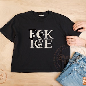 May include: Black t-shirt with the text "FCK ICE" in a distressed white font. The design includes a star, crescent moons, and a flame. The shirt is paired with blue jeans.