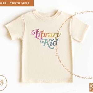 May include: A cream colored t-shirt with the text "Library Kid" in rainbow colors. The text is in a playful font. A portion of every sale is donated to causes we fight for.