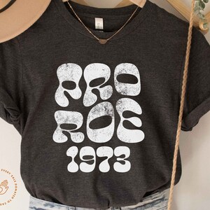 May include: A dark gray t-shirt with a white graphic that says "Pro Roe 1973" in a retro font.