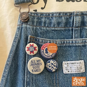 Puede incluir: Peto de mezclilla azul con una etiqueta de Levi Strauss & Co. y cuatro chapas redondas. Las chapas muestran el texto VOTE, VOTE BLOCKHEADS OUT, ALL MY HOMIES VOTE EARLY y vote democracy, con un patrón de estrella en una de ellas.