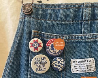 May include: Blue denim overalls featuring a Levi Strauss & Co. patch and four circular pinback buttons. The buttons display the text VOTE, VOTE BLOCKHEADS OUT, ALL MY HOMIES VOTE EARLY, and vote democracy, with a star quilt pattern on one button.