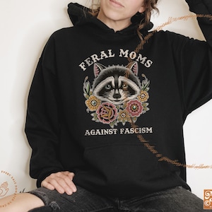 May include: Black hooded sweatshirt with a graphic of a raccoon with flowers and the text "Feral Moms Against Fascism".