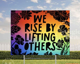 May include: A two-sided yard sign with the text "WE RISE BY LIFTING OTHERS" in black against a rainbow gradient background. The sign is 24 x 18 inches and features black floral silhouettes. Includes a metal stand.
