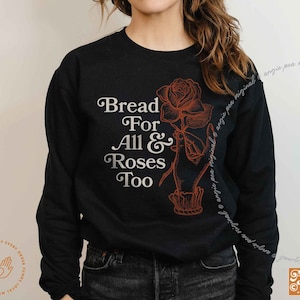 May include: Black long-sleeved sweatshirt with the text "Bread For All & Roses Too" in a vintage style font. A red rose with a stem and leaves is drawn next to the text, with a hand holding the rose.