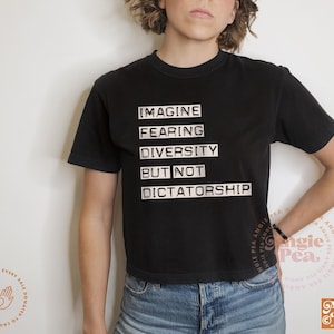 May include: Black t-shirt with the text "IMAGINE FEARING DIVERSITY BUT NOT DICTATORSHIP" in white block letters. The shirt is paired with blue denim jeans. The image includes the Angie Pea logo.