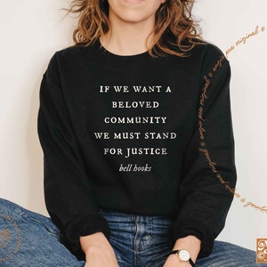 May include: Black sweatshirt with the text "IF WE WANT A BELOVED COMMUNITY WE MUST STAND FOR JUSTICE bell hooks" in white. The sweatshirt has long sleeves and a crew neck. The brand name "Angie Pea" is printed at the bottom right.
