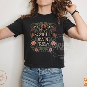 May include: Black t-shirt with the text "FREE SOCIETIES DISSENT FREELY" in a floral design. The design includes flowers and leaves in shades of orange, pink, and green. The shirt is worn with blue jeans.