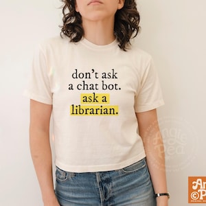 May include: A cream-colored short-sleeve t-shirt featuring the black text "don't ask a chat bot." followed by the text "ask a librarian." highlighted with a yellow rectangular background. The shirt has a crew neckline and a standard fit.