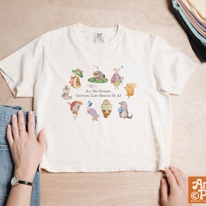 May include: Cream-colored t-shirt featuring a whimsical illustration of various animals, including a cat, squirrel, and owl, arranged in a circle. The text "All My Homies Getting Left Behind By AI" is printed below the illustration. The shirt is paired with blue jeans.