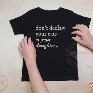 May include: Black t-shirt with the text "don't declaw your cats or your daughters." The shirt has a crew neck and short sleeves. The Angie Pea logo is visible on the bottom right.