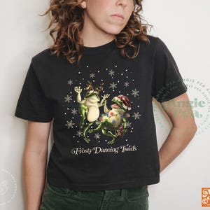 May include: Black t-shirt featuring a graphic of two green frogs dancing with snowflakes. One frog wears a Santa hat, and the other wears antlers. The text "Frosty Dancing Toads" is below the graphic.