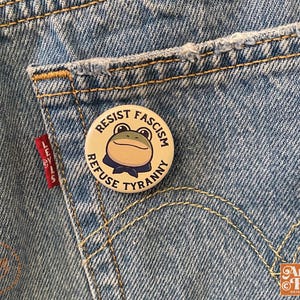 May include: A round button with the text "RESIST FASCISM REFUSE TYRANNY" and a cartoon frog wearing a blue bow tie, pinned to a light blue denim pocket. The button is cream-colored with black text.