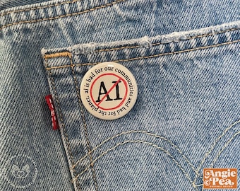 Könnte beinhalten: Ein runder Button mit dem Text "ai is bad for our communities and bad for the planet", der einen roten Kreis mit einem Strich durch die Buchstaben "AI" umgibt. Der Button ist an einer hellblauen Jeans-Hosentasche befestigt.