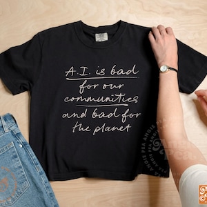 May include: Black t-shirt with the text "A.I. is bad for our communities and bad for the planet" in white script. Light blue denim jeans are visible on the left. A person's arm is on the right.