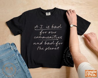 May include: Black t-shirt with the text "A.I. is bad for our communities and bad for the planet" in white script. Light blue denim jeans are visible on the left. A person's arm is on the right.