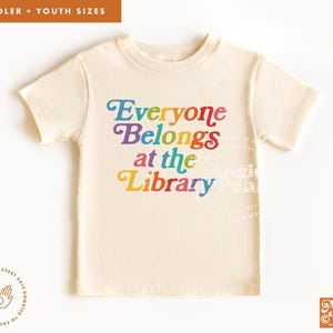 May include: Cream-colored toddler t-shirt with the text "Everyone Belongs at the Library" in rainbow colors. The shirt has short sleeves and a crew neck. The design is centered on the front of the shirt.