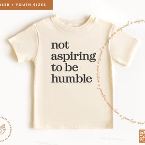 May include: A beige toddler or youth sized t-shirt with the text "not aspiring to be humble" printed in black.