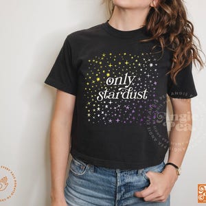 May include: Black t-shirt with the words "only stardust" in white script. The shirt features a gradient of yellow and purple stars. The shirt is paired with blue jeans.