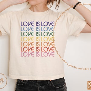 May include: A white t-shirt with the phrase "Love is Love" repeated in rainbow colors. The shirt is cropped and has short sleeves.