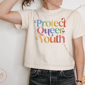 May include: Cream-colored t-shirt with the words "Protect Queer Youth" in rainbow colors. The shirt has a vintage, retro style. The brand name "Angie Pea" is visible in the bottom right corner.