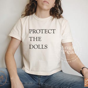 May include: A white cropped t-shirt with the text "PROTECT THE DOLLS" printed in black.