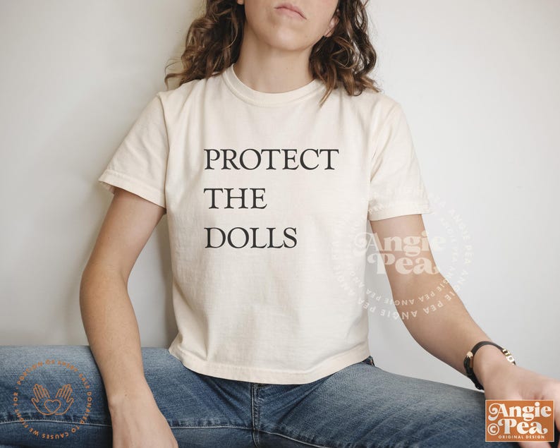 May include: A white cropped t-shirt with the text "PROTECT THE DOLLS" printed in black.