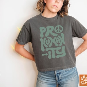 May include: Dark gray t-shirt with the words "PRO HUMANITY" in a teal, retro font. The shirt has a peace sign incorporated into the design. The model is wearing blue jeans. The shirt is from Angie Pea.