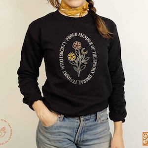 K&ouml;nnte beinhalten: Schwarzes Langarmshirt mit einem runden Grafikdesign mit einem floralen Motiv, einem zunehmenden Mond und dem Text "Proud Member of the Spooky Liberal Feminist Witch Society".