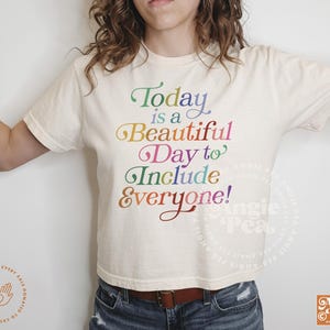 May include: A white cropped t-shirt with the text "Today is a Beautiful Day to Include Everyone!" printed in rainbow colors.
