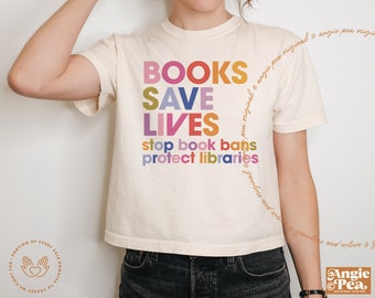 May include: Cream-colored t-shirt with the text "BOOKS SAVE LIVES" in colorful block letters. Below, the shirt reads "stop book bans protect libraries." The shirt has short sleeves and a crew neck.