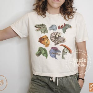 May include: A white t-shirt with a colorful dinosaur graphic print. The dinosaurs are in various poses and colors, including green, orange, blue, and red. The shirt has a crew neck and short sleeves.