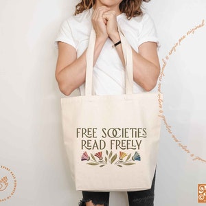 Pode incluir: Uma bolsa de tecido branca com o texto "FREE SOCIETIES READ FREELY" em letras marrons. O texto &eacute; rodeado por um desenho floral com flores coloridas e folhas verdes.